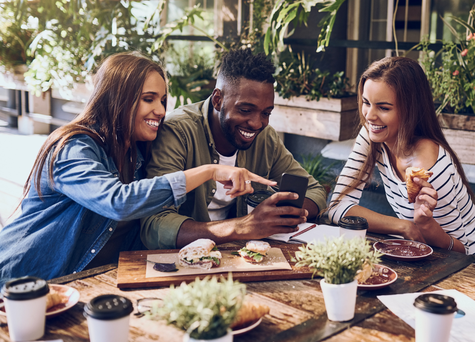 Two women and one man sitting at a table with food with farm chic decor looking at simple checking account on mobile device.