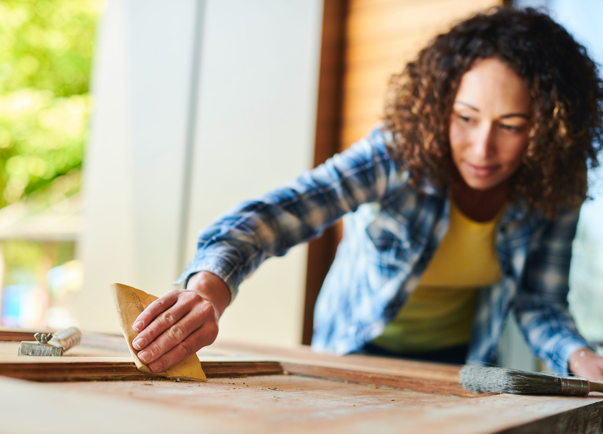 Woman sanding wooden door wearing plaid shirt over yellow shirt.